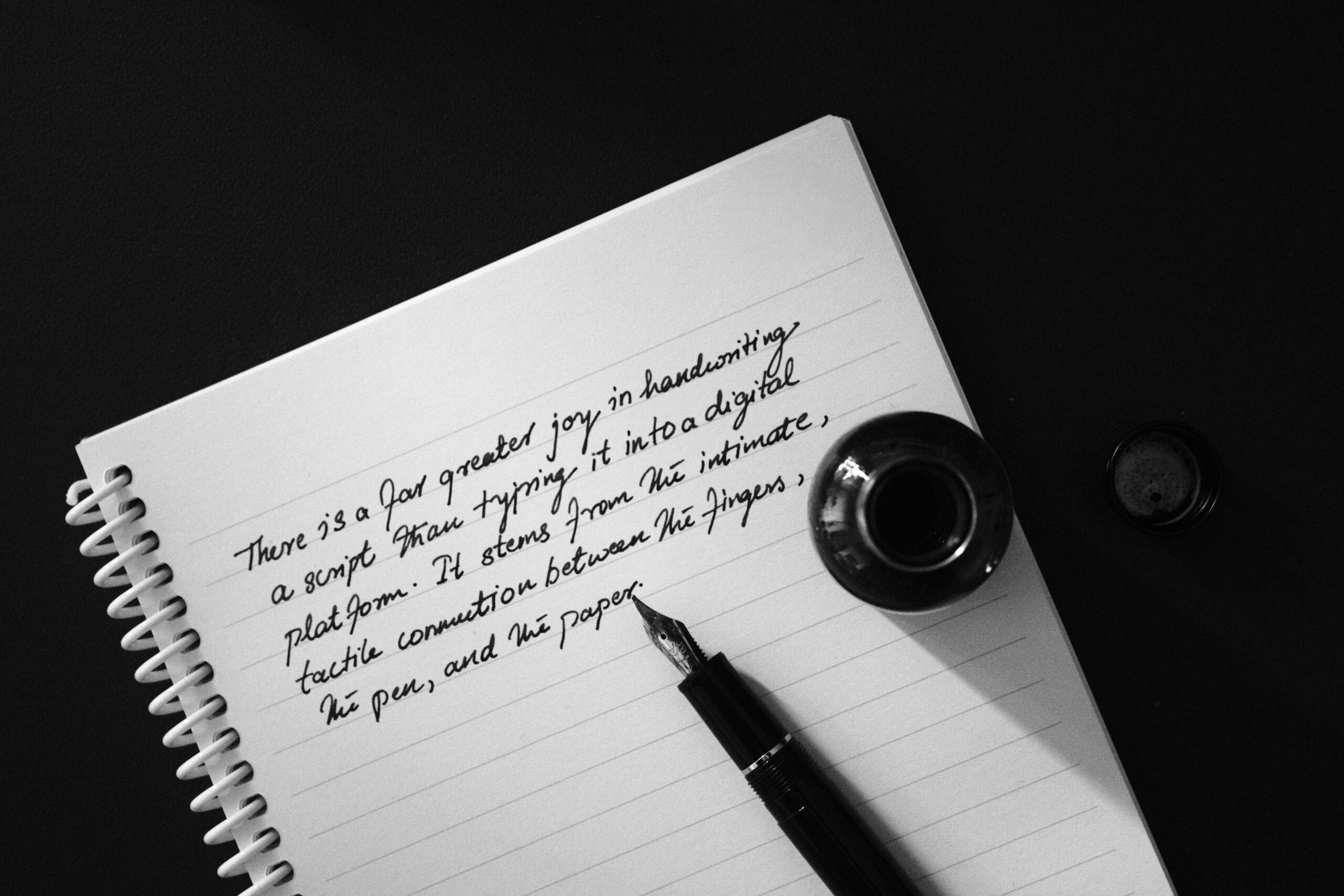 Black and white image of handwritten script with a fountain pen and ink bottle on a notebook.